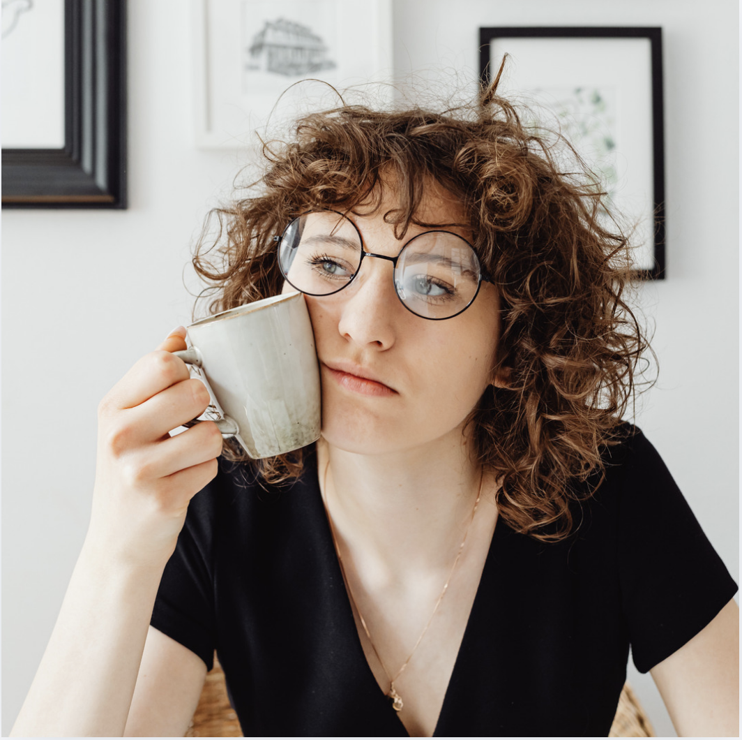 A young woman with curly hair and glasses rests a mug against her cheek, looking thoughtful and reflective.